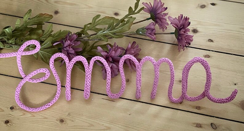 May include: Pink knitted yarn spelling out the name "Emma" on a light wood background with a sprig of purple flowers.