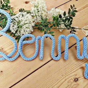 May include: A light blue knitted name sign spelling "Benny" on a wooden surface.