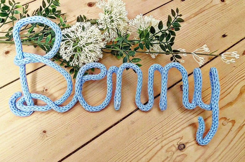May include: A light blue knitted name sign spelling "Benny" on a wooden surface.