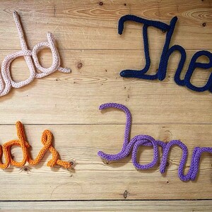 May include: Four knitted names, "Jada", "Theo", "Madis", and "Jonna", are displayed on a wooden surface. The names are made of different colored yarn: pink, blue, orange, and purple.