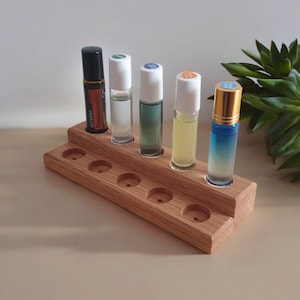 Può includere: Porta bottiglie di olio essenziale in legno con otto fessure. Il supporto è in legno marrone chiaro e ha una finitura naturale. Sei bottiglie di vetro trasparente con liquidi di diversi colori sono esposte nel supporto. Una bottiglia ha un'etichetta marrone con il testo "dōTERRA Frankincense".