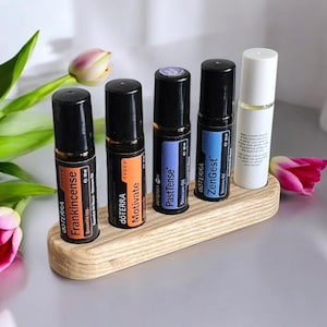 Puede incluir: Un soporte de madera para botellas de rodillo con cinco ranuras, que contiene cinco botellas de aceite esencial de rodillo diferentes. Las botellas están etiquetadas como "Frankincense", "Motivate", "Past Tense", "ZenGest" y "dōTERRA".