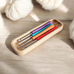 May include: A wooden tray containing four colourful crochet hooks. The hooks are arranged in a row, with the handles facing upwards. The tray is made of light wood and has a rectangular shape.