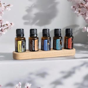 Puede incluir: Un soporte de madera para aceites esenciales con seis ranuras, cada una con una pequeña botella de vidrio ámbar con una etiqueta blanca. Las etiquetas tienen el nombre de la marca "dōTERRA" y el nombre del aceite, como "Lemon", "Tangerine", "Peppermint", "AromaTouch" y "On Guard".