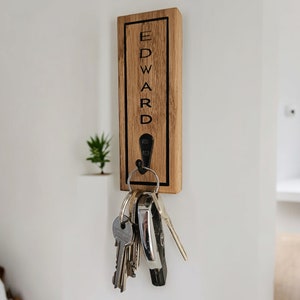 May include: Wooden key holder with the name "EDWARD" engraved on it. The holder has a black metal hook with a set of keys hanging from it.
