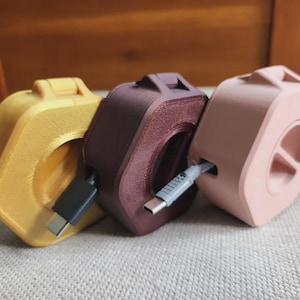 Può includere: Tre organizzatori di cavi esagonali in giallo, viola scuro e rosa chiaro. Ogni dispositivo in plastica presenta un'apertura centrale e una fessura laterale per avvolgere i cavi di ricarica, con connettori USB-C che sporgono dalle unità gialla e viola.