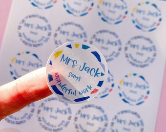 May include: Sheet of transparent stickers with a blue border and colorful geometric shapes. Each sticker says "Mrs Jack says wonderful work" in a handwritten font. The stickers are on a white background with a pink surface.