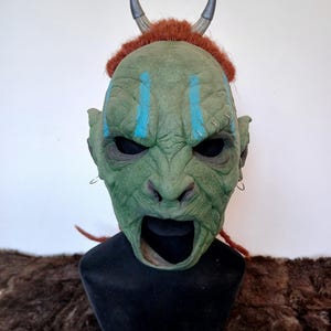 Può includere: Maschera dettagliata verde e blu con corna e capelli rossi. La maschera ha una bocca spalancata e una superficie testurizzata.