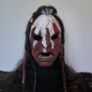 Può includere: Una maschera dipinta di rosso e bianco con un'espressione feroce. La maschera ha una lunga treccia nera con perline rosse e marroni.