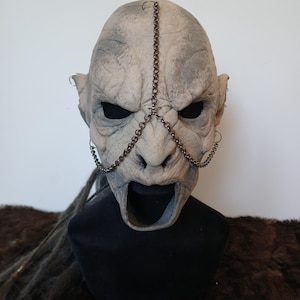 Può includere: Una maschera in lattice grigia e bianca con una catena sul viso e una grande bocca aperta. La maschera è su un supporto nero con uno sfondo di pelliccia marrone.