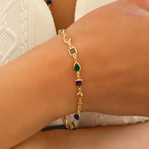 Könnte beinhalten: Ein goldfarbenes Halsketten- und Armband-Set. Die Halskette weist eine Reihe farbiger Edelsteine in verschiedenen Formen auf, darunter rund, quadratisch und tropfenförmig. Das Armband spiegelt das Design der Halskette wider, mit ähnlichen Edelsteinen.