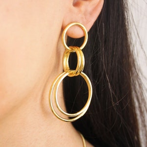 May include: Gold hoop earrings with multiple interlocking circles. The earrings are large and dangle from the earlobe.