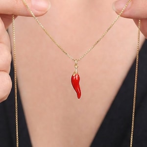 Puede incluir: Un collar dorado con un pequeño colgante de pimiento rojo. El colgante es de color rojo vibrante y cuelga de una delicada cadena dorada. El collar se muestra sobre un fondo oscuro.