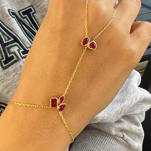 Könnte beinhalten: Ein goldfarbenes Handkettenarmband mit drei roten Edelsteinakzenten. Das Armband verbindet sich mit einem Ring und hat ein filigranes Kettendesign. Die Edelsteine sind tiefrot und in einem goldfarbenen Metall gefasst.