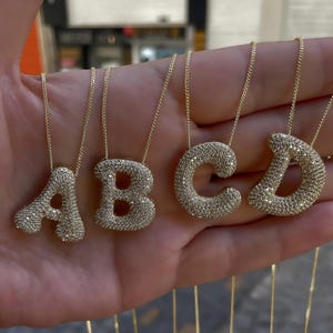 May include: Four gold-colored necklaces with the letters A, B, C, and D as pendants. Each letter is encrusted with small, sparkling stones. The necklaces are displayed against a neutral background.