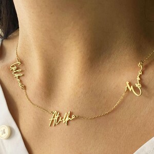 Op de afbeelding: Een goudkleurige ketting met drie gepersonaliseerde naamhangers in een cursief lettertype. De ketting wordt om de nek van een persoon gedragen, met de namen langs de ketting gerangschikt.