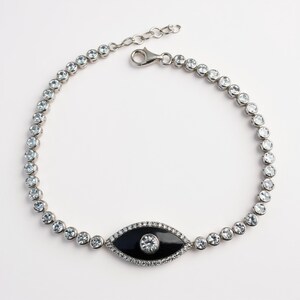 May include: A silver bracelet featuring a series of clear, round gemstones. The bracelet includes a central black enamel eye-shaped charm with a central clear gemstone and surrounding clear gemstones. The bracelet has a lobster clasp closure.