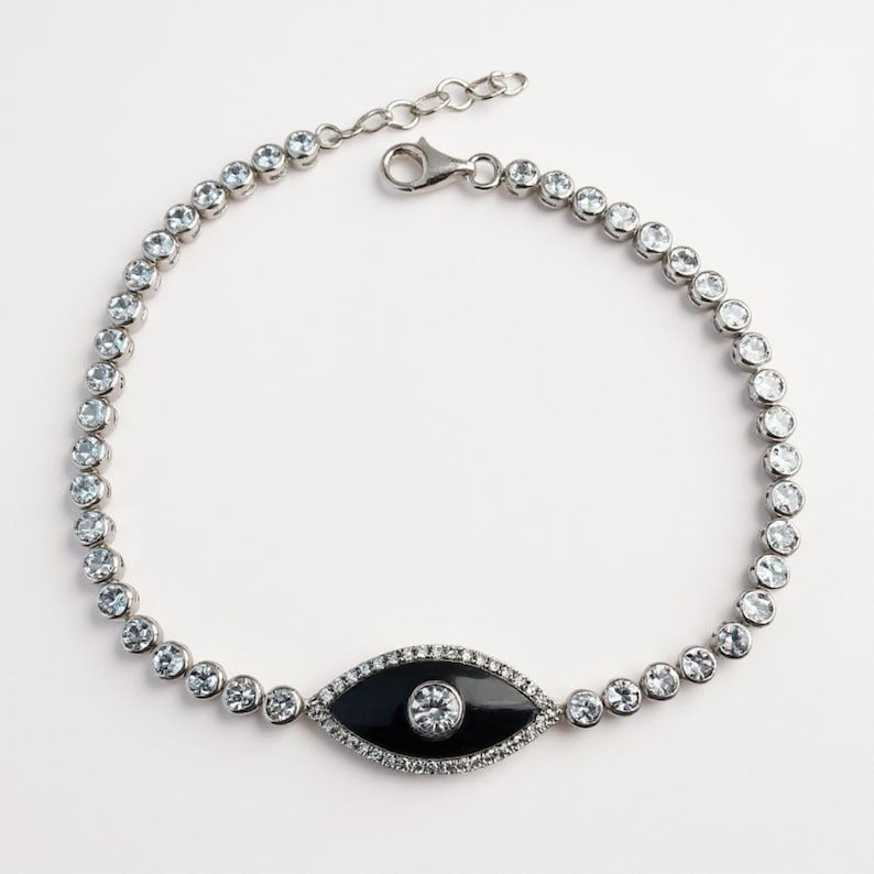 May include: A silver bracelet featuring a series of clear, round gemstones. The bracelet includes a central black enamel eye-shaped charm with a central clear gemstone and surrounding clear gemstones. The bracelet has a lobster clasp closure.