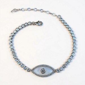 May include: Silver-toned bracelet featuring a central eye-shaped charm with a white background and a central clear stone. The bracelet is adorned with multiple round, clear stones. The clasp is a lobster claw style.
