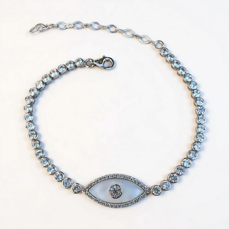 May include: Silver-toned bracelet featuring a central eye-shaped charm with a white background and a central clear stone. The bracelet is adorned with multiple round, clear stones. The clasp is a lobster claw style.