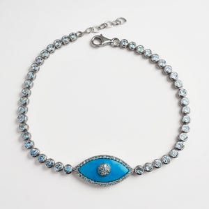 May include: Silver-toned bracelet featuring a turquoise eye-shaped charm with a central diamond-like accent. The bracelet is adorned with round, light blue gemstones. The clasp is a lobster claw style.