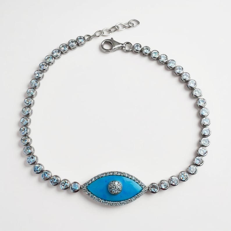 May include: Silver-toned bracelet featuring a turquoise eye-shaped charm with a central diamond-like accent. The bracelet is adorned with round, light blue gemstones. The clasp is a lobster claw style.