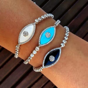 May include: Three silver bracelets with eye-shaped pendants. Each pendant features a different colored center: white, turquoise, and black. The pendants are outlined with small, clear stones, and the bracelets have a delicate, linked design.