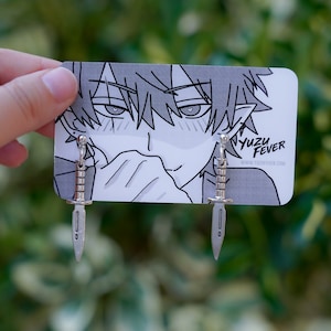 Puede incluir: Pendientes de daga plateados. Los pendientes se muestran en una tarjeta con una ilustración en blanco y negro de estilo anime y el texto "YUZU FEVER". Los pendientes tienen un mango detallado y una hoja puntiaguda.