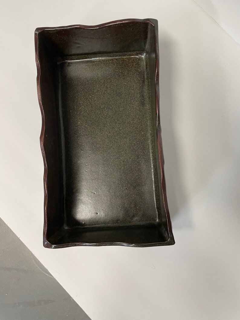 May include: A rectangular ceramic dish with a dark green glaze and a brown rim. The dish has an irregular, wavy edge.