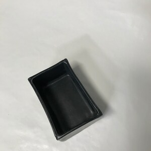 May include: A black rectangular ceramic planter with a slightly curved rim. The planter is empty and is sitting on a white surface.