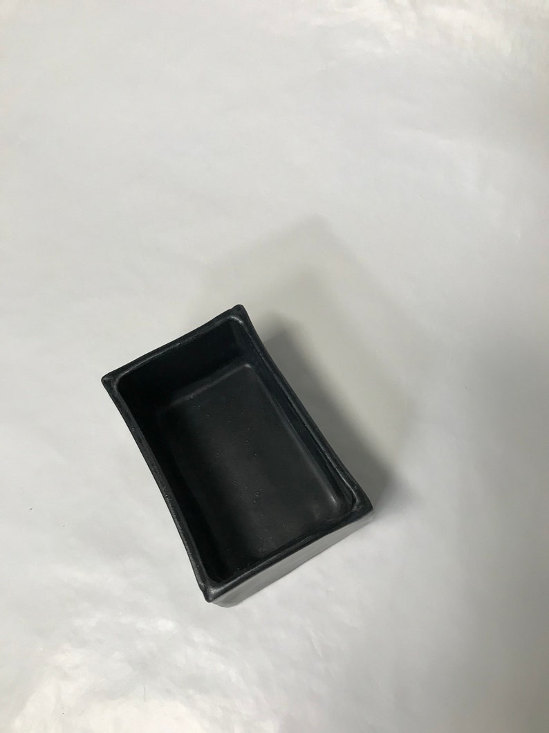 May include: A black rectangular ceramic planter with a slightly curved rim. The planter is empty and is sitting on a white surface.