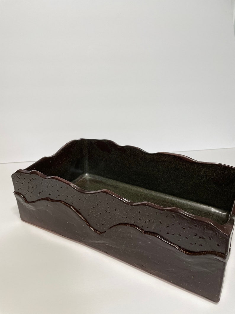 May include: A rectangular ceramic planter with a dark brown glaze and a wavy, textured rim. The planter has a dark green interior.