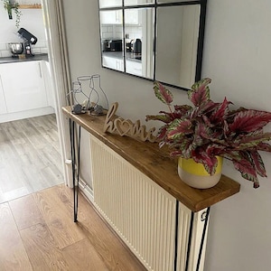 May include: A rustic wooden console table with black hairpin legs sits on top of a white radiator. The table is decorated with a wooden sign that says "home" with a heart in the middle, two black vases, and a potted plant with red and green leaves.
