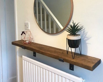 May include: A rustic wooden shelf with metal brackets, mounted above a white radiator. The shelf is decorated with a geometric candle holder, a potted succulent plant, and a round mirror with a copper frame.