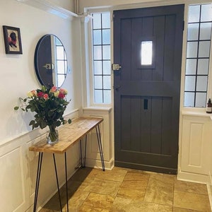 May include: A wooden console table with black hairpin legs, a round mirror, and a bouquet of pink roses in a glass vase. The table is in a hallway with a dark grey door and two windows with frosted glass.