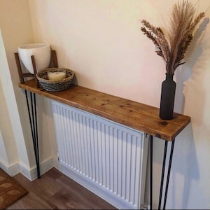 May include: A rustic wooden console table with black hairpin legs. The table is placed over a white radiator and decorated with a white planter, a wicker basket, and a vase with dried flowers.
