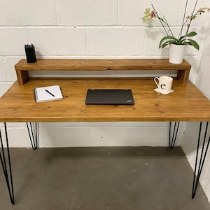 May include: A rustic wooden desk with a shelf, black hairpin legs, and a laptop computer on the surface. A notebook, pen, and a white mug with the letter "e" are also on the desk.