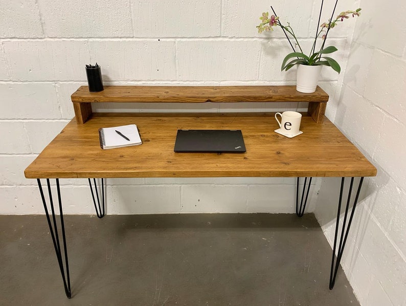 May include: A rustic wooden desk with a shelf, black hairpin legs, and a laptop computer on the surface. A notebook, pen, and a white mug with the letter "e" are also on the desk.