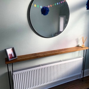 May include: A wooden console table with black metal legs. The table is placed in front of a white radiator and has a framed photo on top. A round mirror with a black frame hangs on the wall above the table. A colorful garland of paper circles hangs above the mirror.