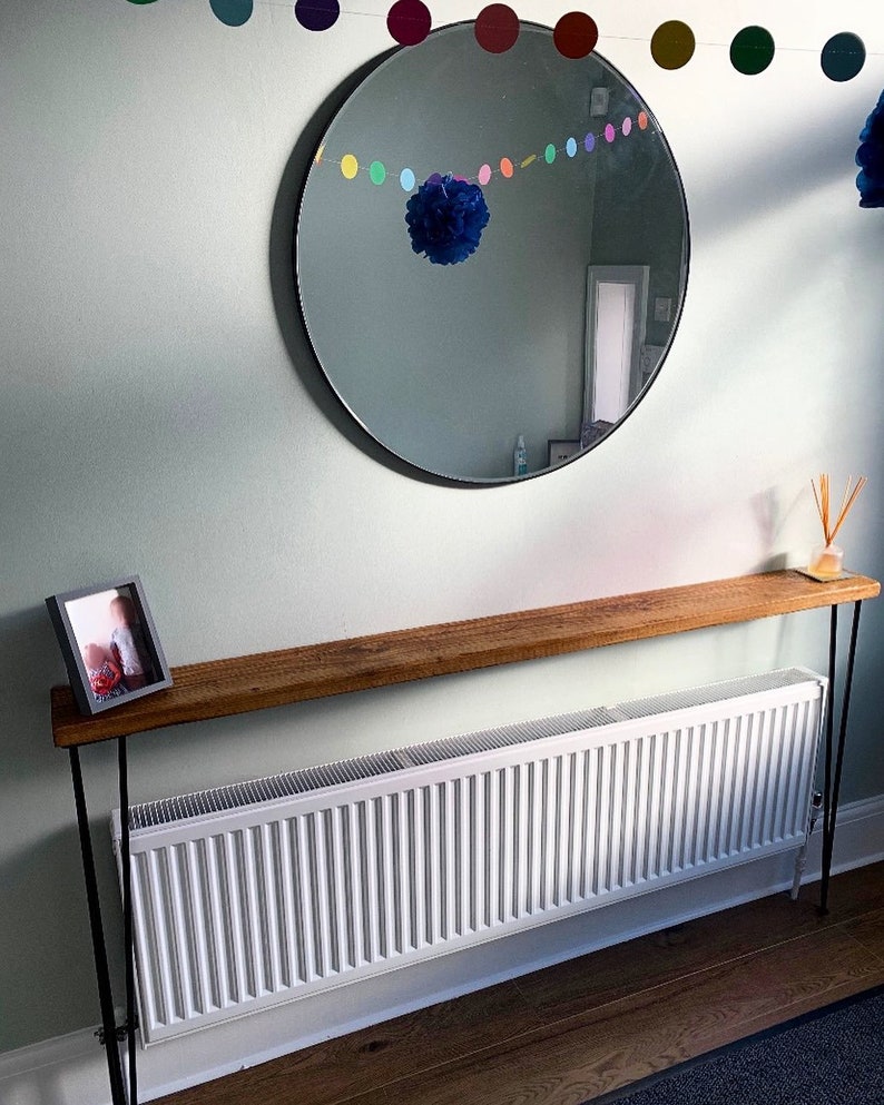 May include: A wooden console table with black metal legs. The table is placed in front of a white radiator and has a framed photo on top. A round mirror with a black frame hangs on the wall above the table. A colorful garland of paper circles hangs above the mirror.