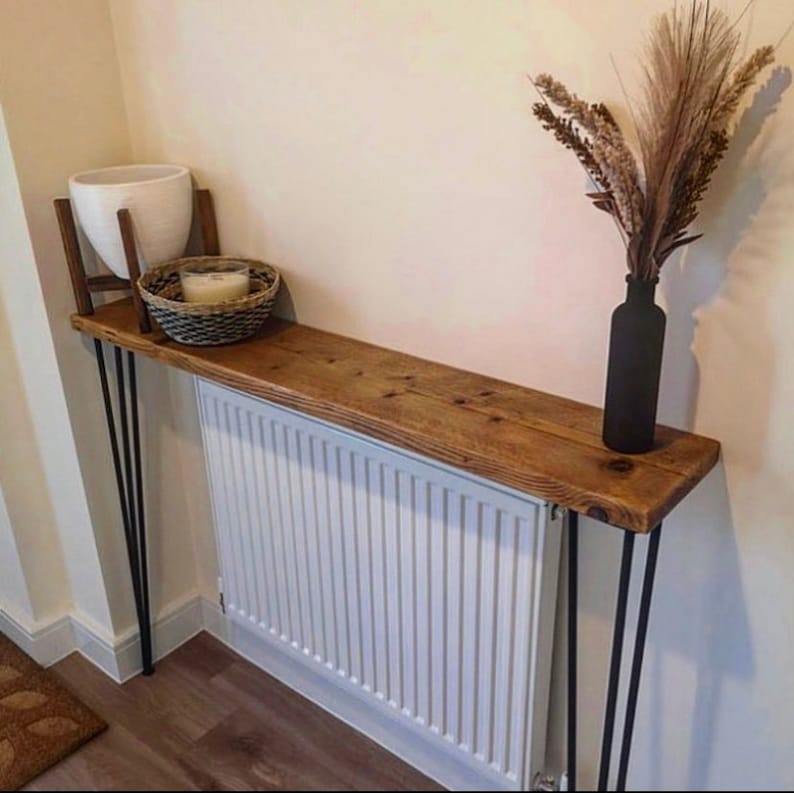 May include: A rustic wooden console table with black hairpin legs. The table is placed over a white radiator and decorated with a white planter, a wicker basket, and a vase with dried flowers.