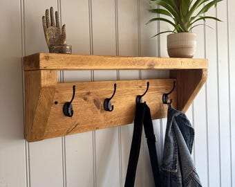 May include: A wooden wall shelf with four black metal hooks. A denim jacket and a black bag hang from the hooks. A decorative hand statue and a potted plant with a red flower sit on the shelf. The shelf is made of light brown wood.