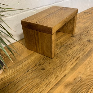Puede incluir: Un peque&ntilde;o taburete de madera con un acabado de madera natural. El taburete est&aacute; hecho de madera reciclada y tiene un aspecto r&uacute;stico.