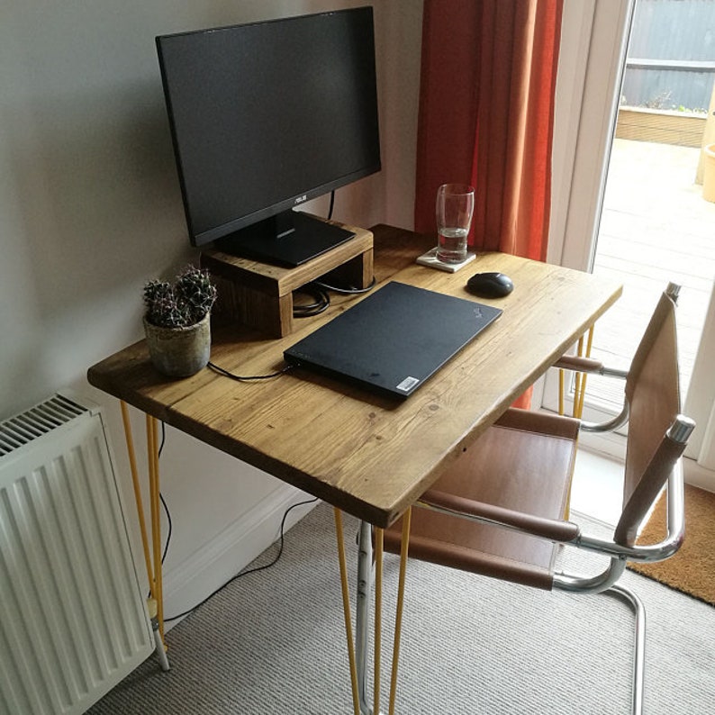 Puede incluir: Un escritorio de madera con un acabado marr&oacute;n claro y patas de horquilla amarillas. Un ordenador port&aacute;til, un monitor y un cactus est&aacute;n sobre el escritorio. Una silla de cuero marr&oacute;n con un marco de cromo est&aacute; al lado del escritorio.