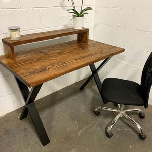 May include: A rustic wooden desk with a black metal X-shaped base. The desk has a shelf above the desktop. A black office chair with a mesh back is positioned next to the desk.