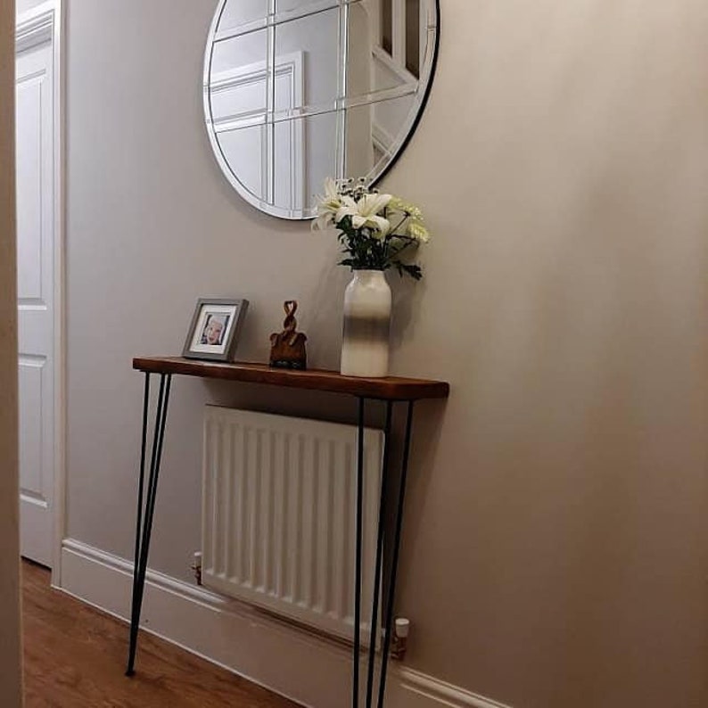 May include: A wooden console table with black hairpin legs. The table is decorated with a vase of white flowers, a framed photo, and a small wooden figurine. The table is in front of a white radiator and a round mirror with a grid pattern.