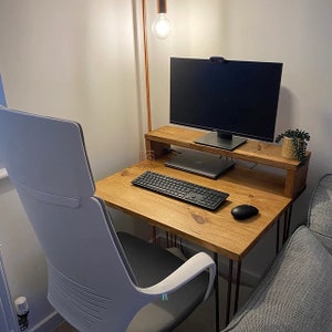 May include: A wooden desk with a light wood finish and black metal legs. The desk has a monitor stand and a keyboard on the surface. A white office chair is positioned in front of the desk.