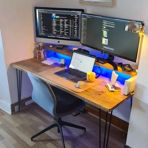 May include: A wooden desk with black hairpin legs, a gray office chair, two computer monitors, a laptop, a yellow mug, and a blue light strip under the desk.
