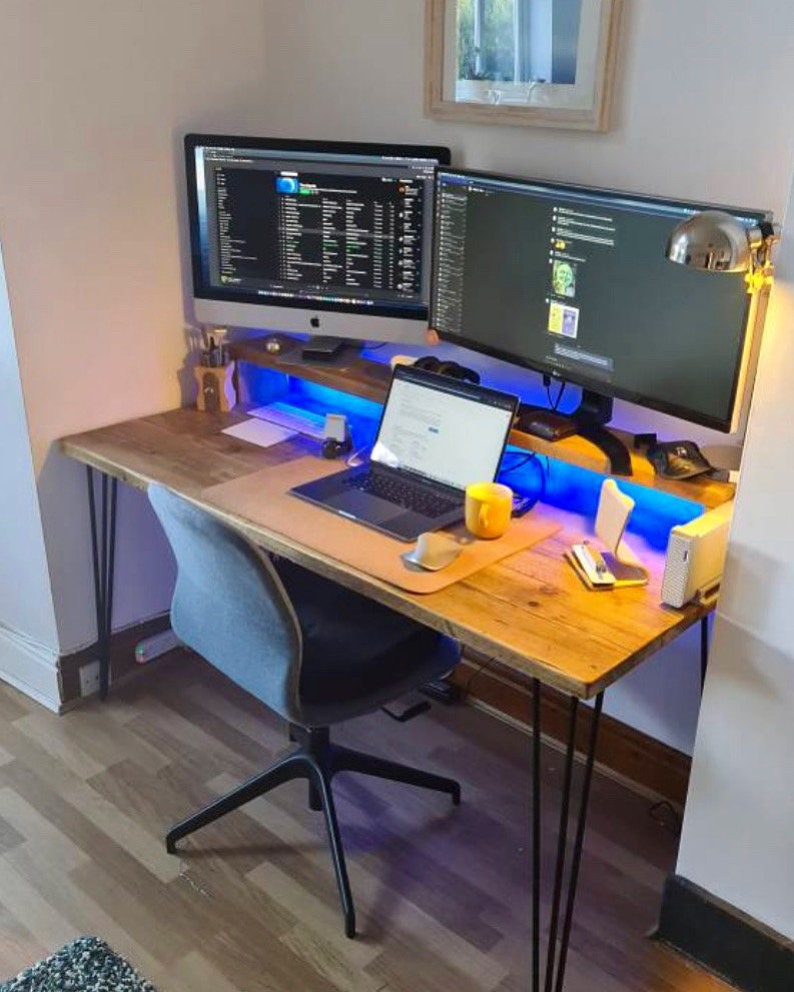 May include: A wooden desk with black hairpin legs, a gray office chair, two computer monitors, a laptop, a yellow mug, and a blue light strip under the desk.