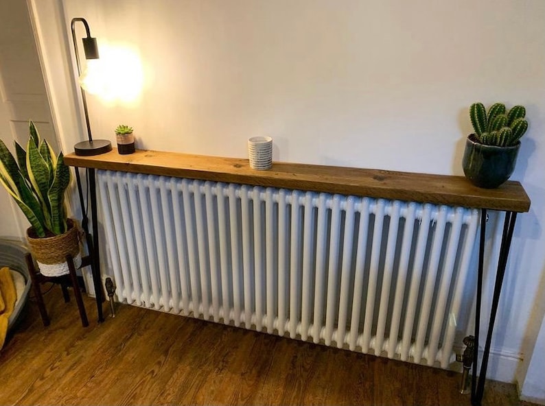 May include: A rustic wooden shelf with black metal hairpin legs sits atop a white radiator. The shelf is decorated with a small potted plant and a lamp.